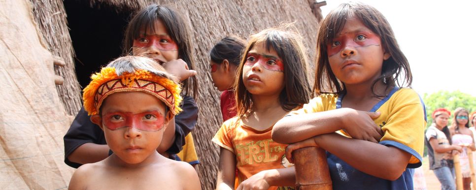 Vier indigene Kinder aus Brasilien stehen draußen vor einer Hütte. Die Augen sind mit einem Band aus roter Farbe umzogen.